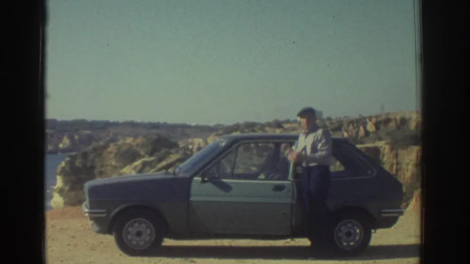 A man stands by an open hatchback door on a coastal dirt road in 1976 Greece home-movie footage.