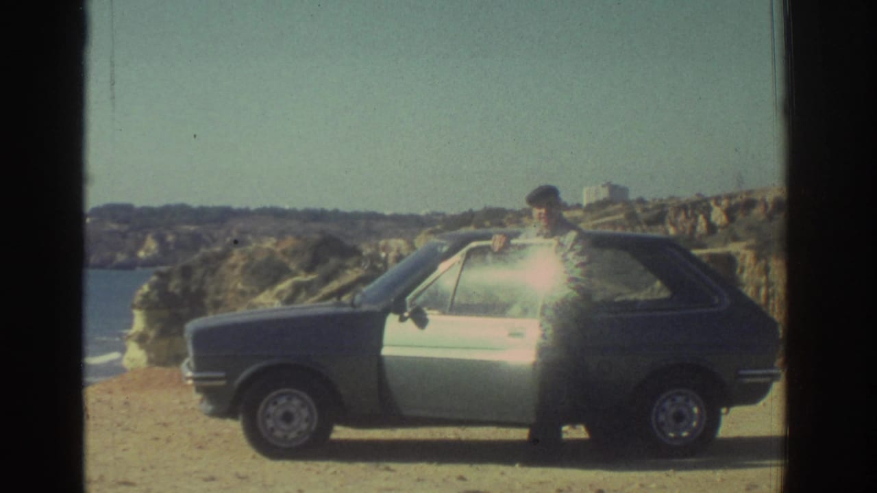 Man in a cap stands beside a blue and white hatchback near coastal cliffs.