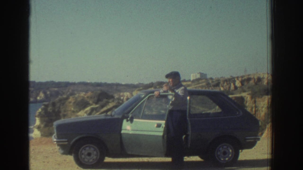A man in a flat cap opens a compact hatchback door on a sandy coastal overlook in Greece, 1976.