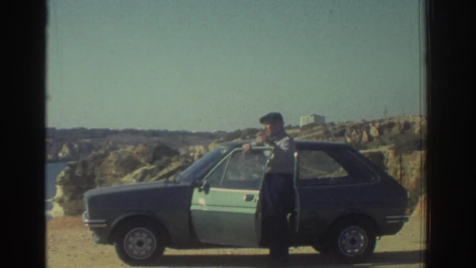 A man in a flat cap opens a compact hatchback door on a sandy coastal overlook in Greece, 1976.