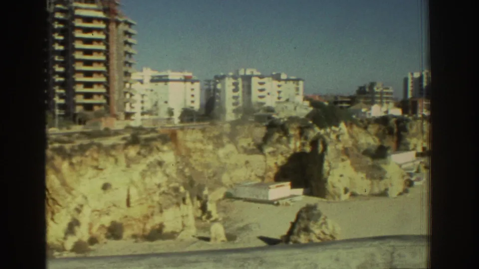 1976 Greece coastal view with pale cliffs, beach below, and apartment blocks rising behind the shoreline.