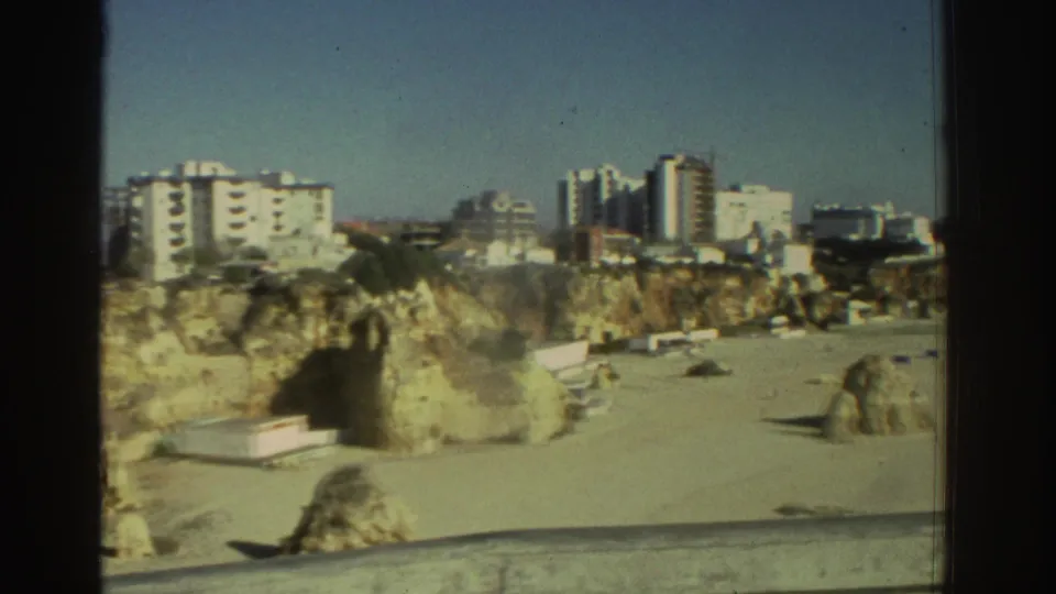 Home-movie view of a Greek coastal cliff, sandy beach, apartment blocks, and distant vehicles in 1976.