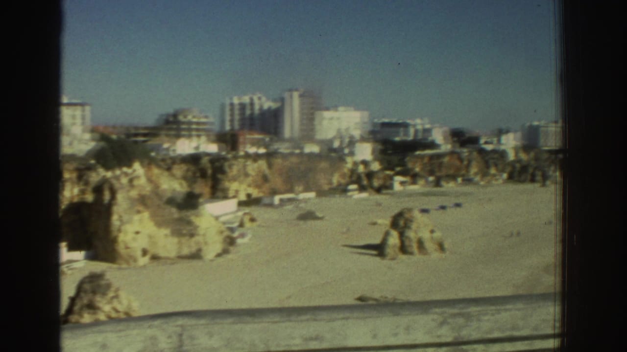 1976 Greece home-movie frame showing a balcony view over a sandy beach, rocky cliff edge, and distant modern buildings.