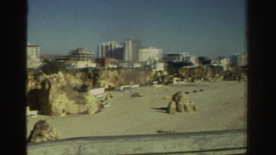 1976 Greece home-movie frame showing a balcony view over a sandy beach, rocky cliff edge, and distant modern buildings.