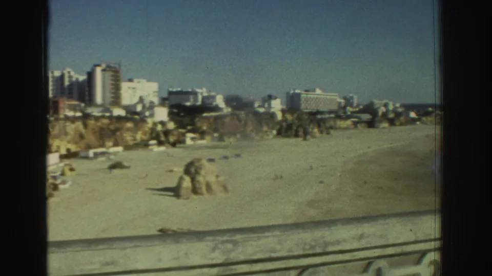1976 Greece coastal view from a balcony, showing the beach, rocky foreground, distant town buildings, and open sky.
