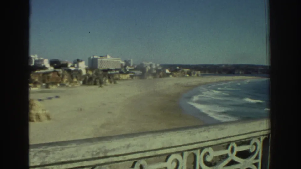 1976 Greece balcony view as the pan opens toward a curving beach, low surf, distant buildings, and coastline.
