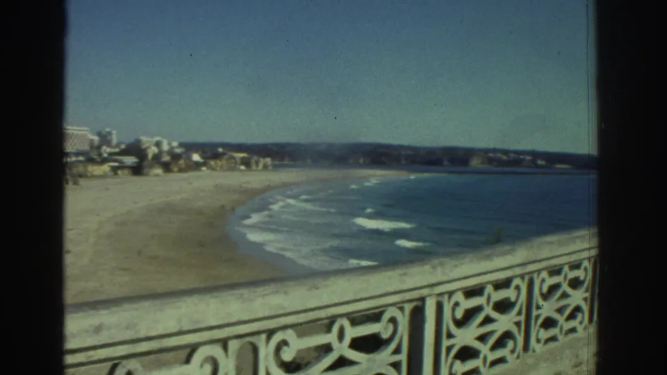 1976 Greece home-movie frame showing an ornate balcony railing above a sweeping beach, sea, and distant coast.