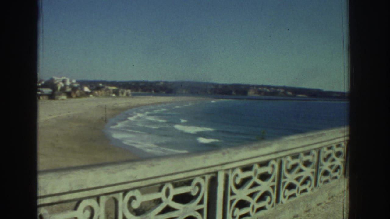 1976 Greece home-movie frame looking past an ornate pier railing toward a quiet beach, small waves, and distant shoreline.