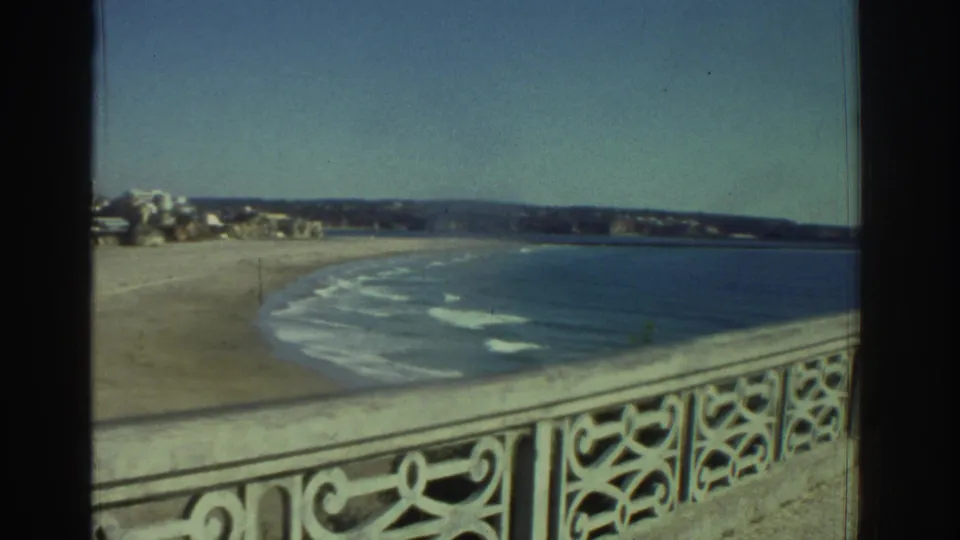 1976 Greece home-movie frame looking past an ornate pier railing toward a quiet beach, small waves, and distant shoreline.