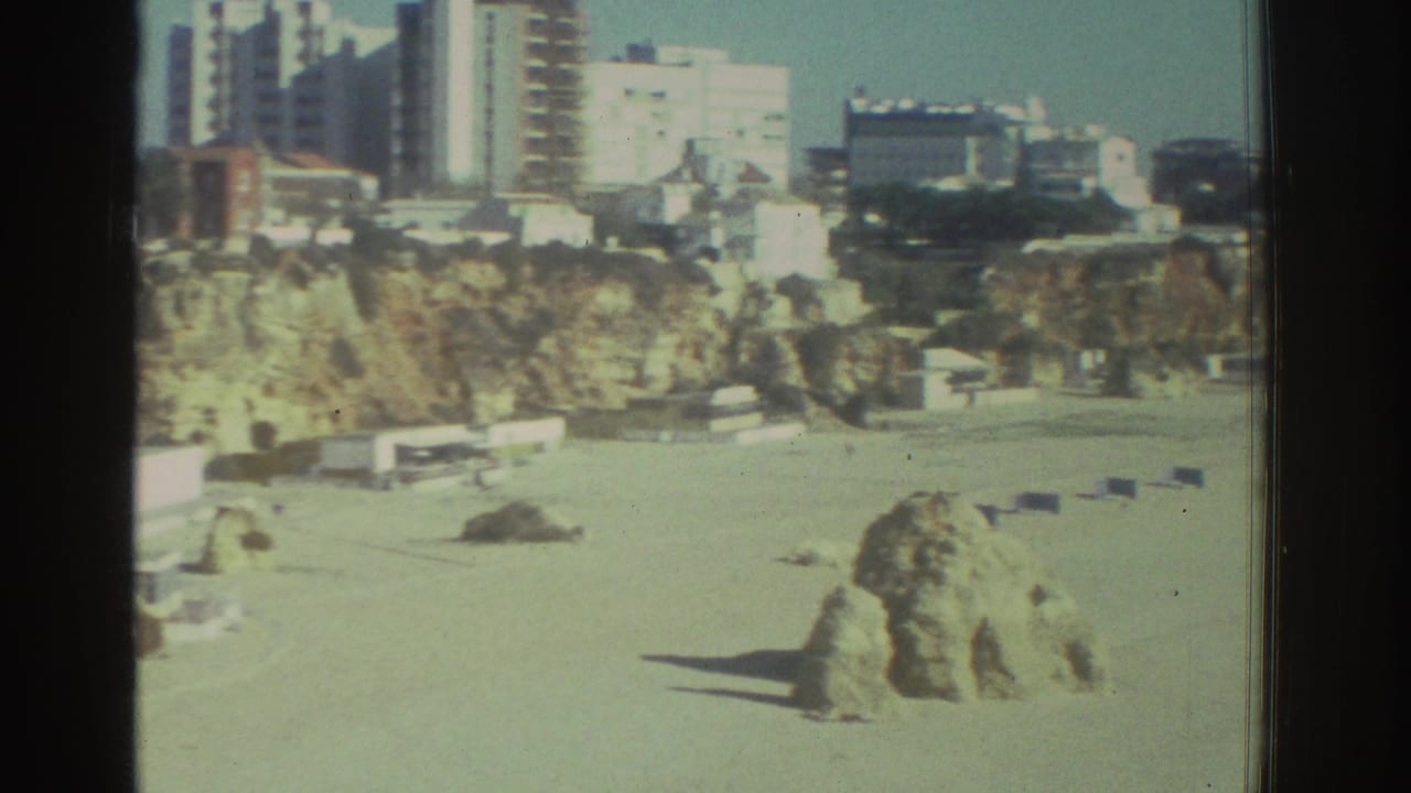 Beach below coastal cliffs with buildings above and small distant figures on the sand.