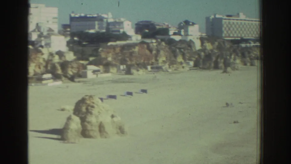 1976 Greece beach frame showing a large rock on the sand, cliffside structures, and a quiet waterfront skyline.