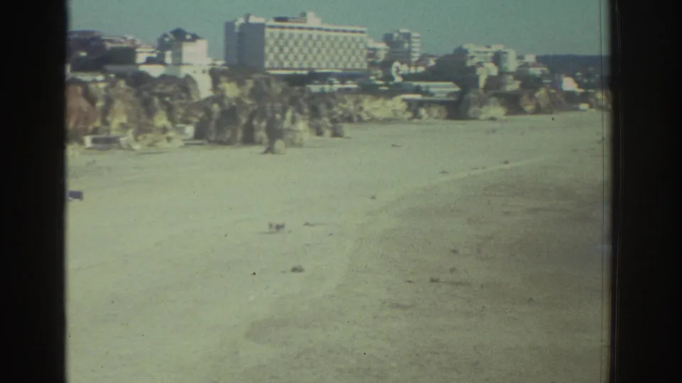 1976 Greece home-movie view across a quiet beach toward cliffside buildings and the shoreline at frame edge.