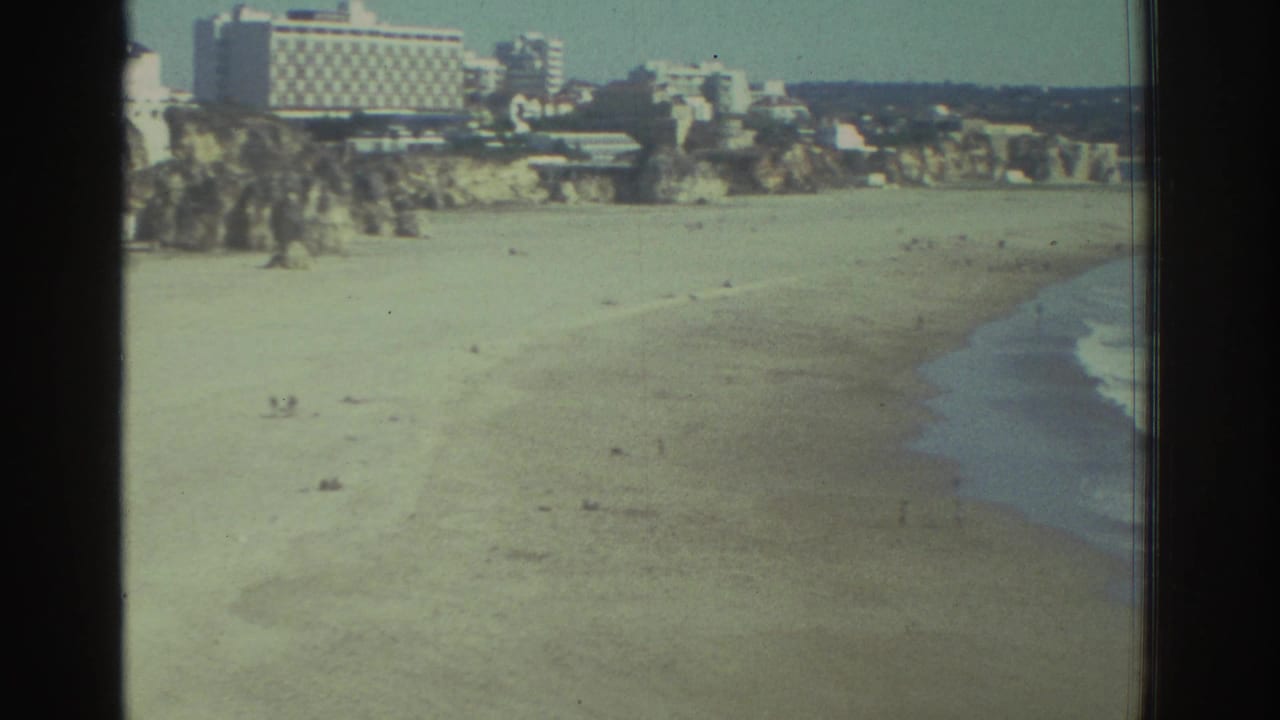 Wide sandy beach with surf, cliffs, and buildings above the shoreline.