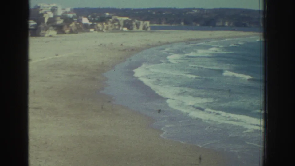 1976 Greece 8mm frame looking along a curving sandy beach with waves and cliffside buildings beyond.