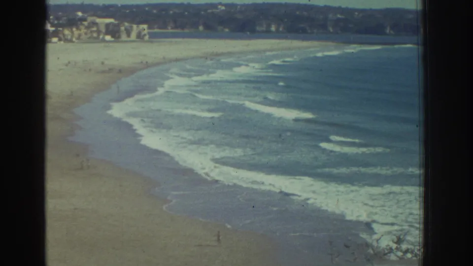 1976 Greece 8mm frame of surf rolling along a sandy beach, with distant beachgoers and buildings beyond.