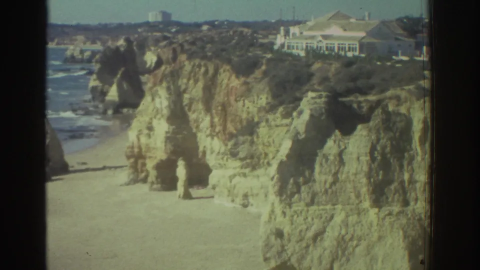 1976 Greece 8mm frame showing rugged seaside cliffs, a beach below, and coastal buildings near the cliff edge.