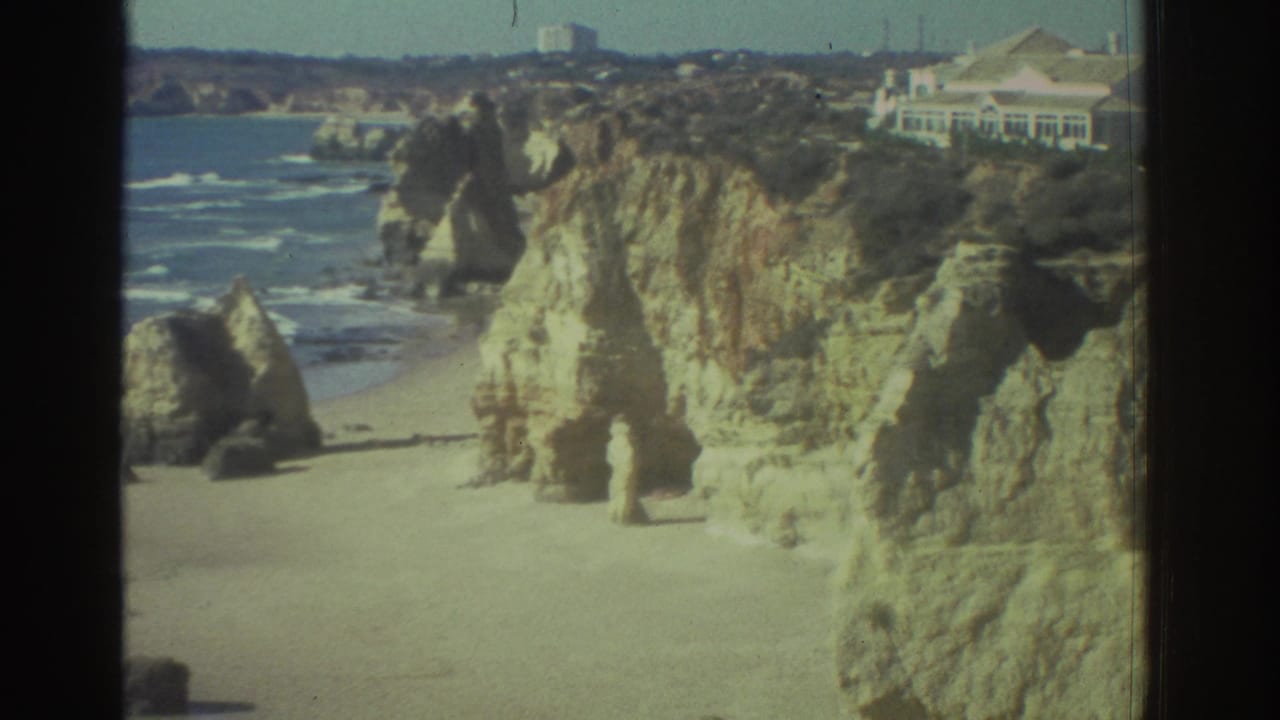 1976 Greece 8mm frame showing a sandy Mediterranean beach, limestone cliffs, surf, and a distant white building on the headland.
