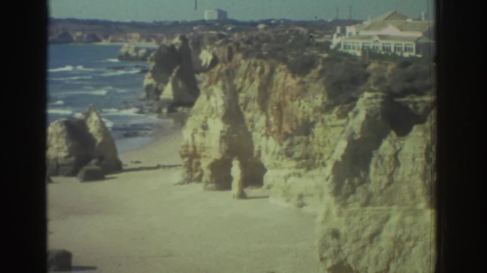 1976 Greece 8mm frame showing a sandy Mediterranean beach, limestone cliffs, surf, and a distant white building on the headland.