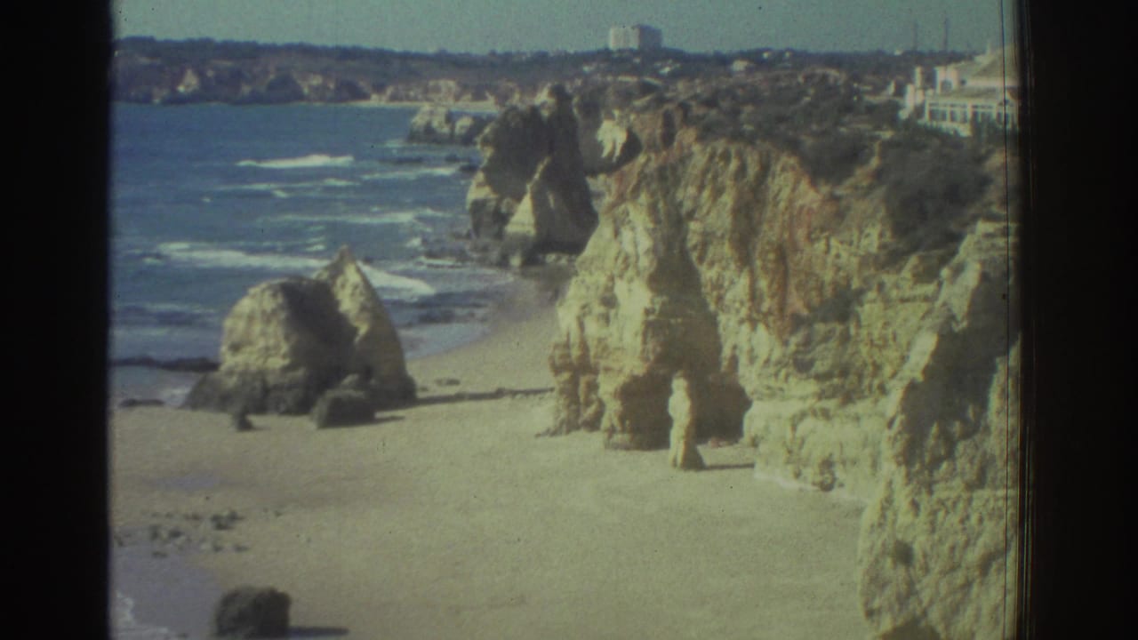 1976 Greece coastal view with pale cliffs, sandy beach, blue surf, and a distant white building near the cliff edge.