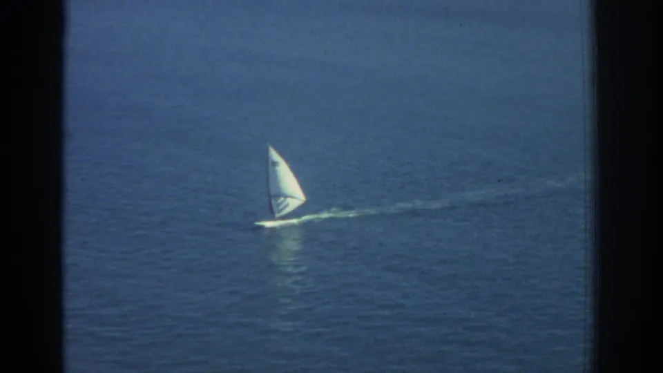 1976 Greece 8mm frame of a lone white-sailed windsurfer moving across deep blue Mediterranean water.