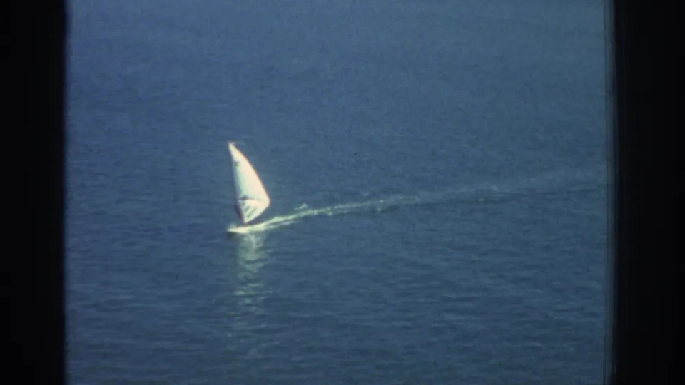 1976 Greece coastal home-movie frame showing a small windsurfer with a white sail crossing calm blue water.