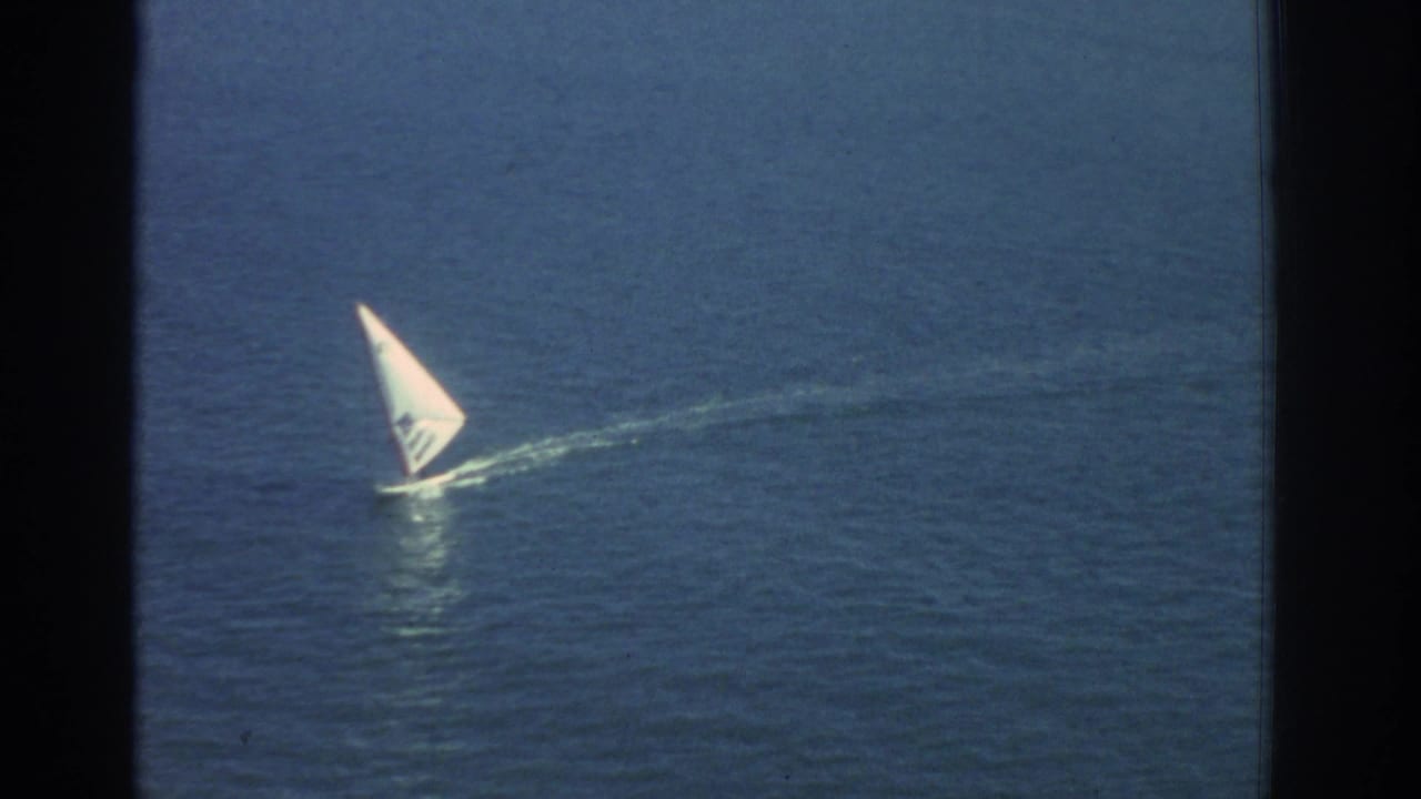 A small white sailboat moves across blue water in 1976 Greece travel footage, seen with soft 8mm grain and dark film edges.