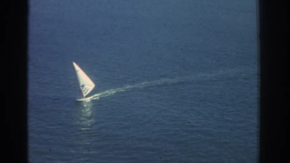 A small white sailboat moves across blue water in 1976 Greece travel footage, seen with soft 8mm grain and dark film edges.