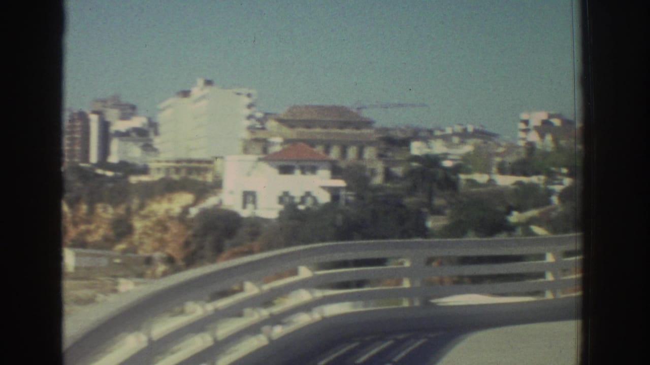 Coastal city buildings in Greece appear beyond a curved bridge railing, with pale cliffs and a clear sky in faded 8mm color.