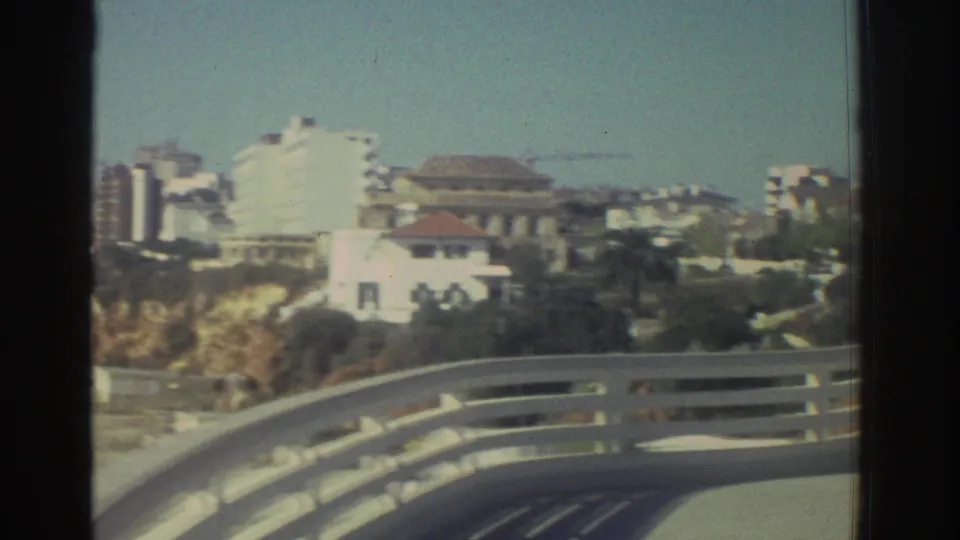Coastal city buildings in Greece appear beyond a curved bridge railing, with pale cliffs and a clear sky in faded 8mm color.