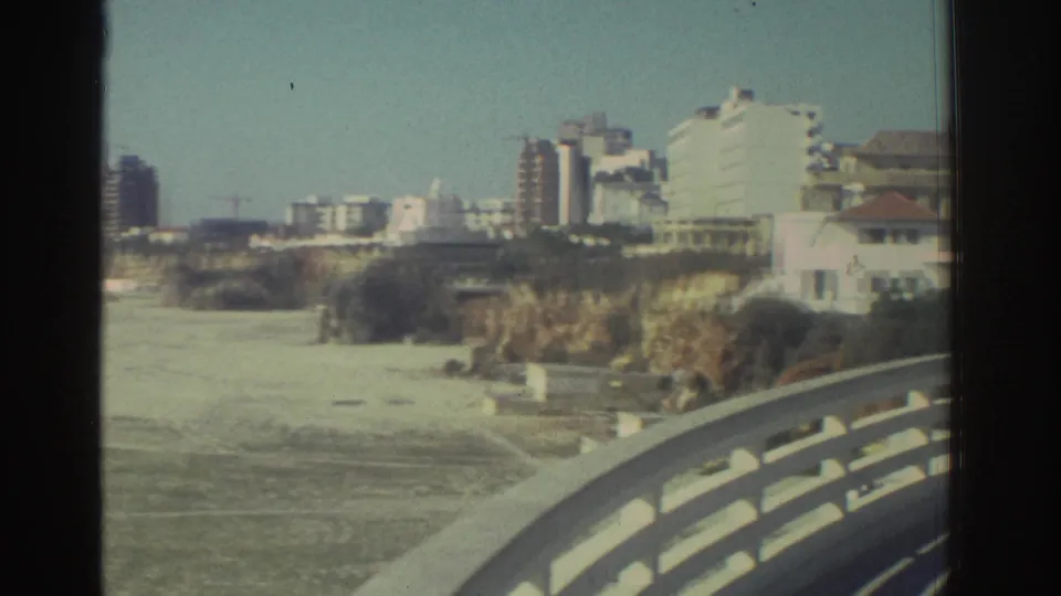 A sandy coastal stretch, cliffs, white buildings, and bridge railing pass through the frame in 1976 Greece home-movie footage.