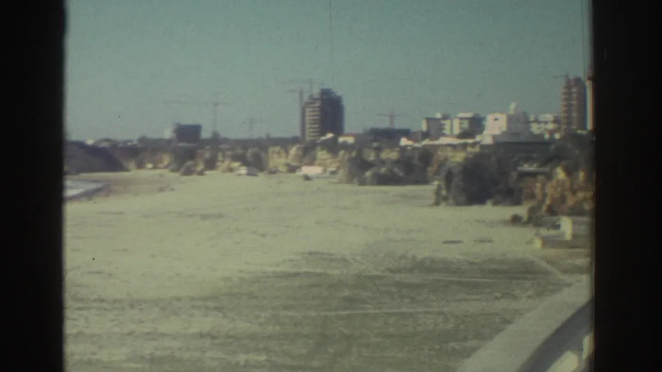 A wider bridge view shows a sandy coastal inlet, distant apartment blocks, cliffs, and construction cranes in 1976 Greece footage.