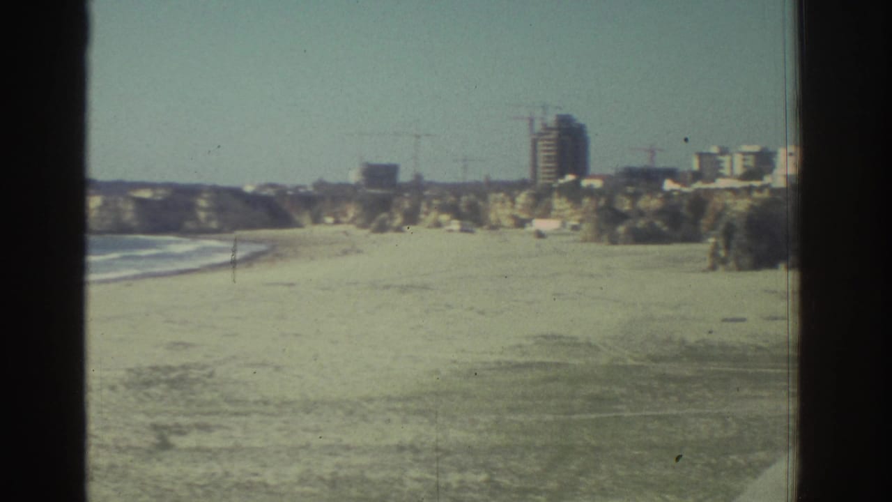 1976 Greece home-movie frame showing a beach, low surf, rocky coastline, and distant construction or buildings on the hillside.