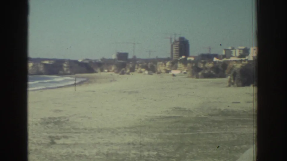 1976 Greece home-movie frame showing a beach, low surf, rocky coastline, and distant construction or buildings on the hillside.
