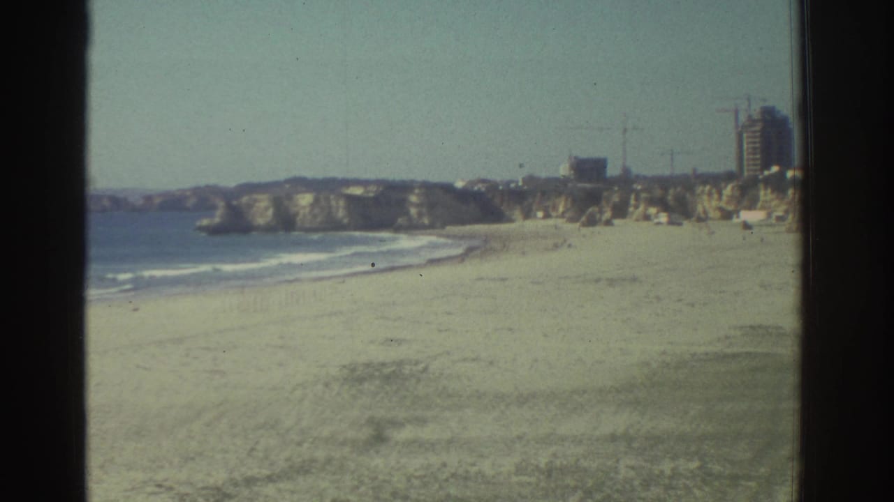 1976 Greece travel footage frame with a sandy beach, Mediterranean surf, rocky cliffs, and distant hillside buildings.