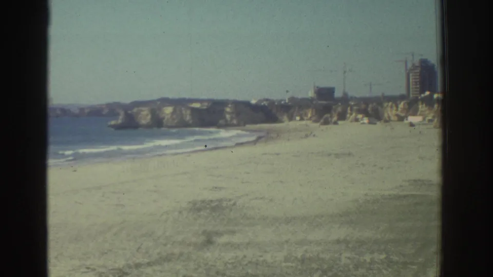 1976 Greece travel footage frame with a sandy beach, Mediterranean surf, rocky cliffs, and distant hillside buildings.