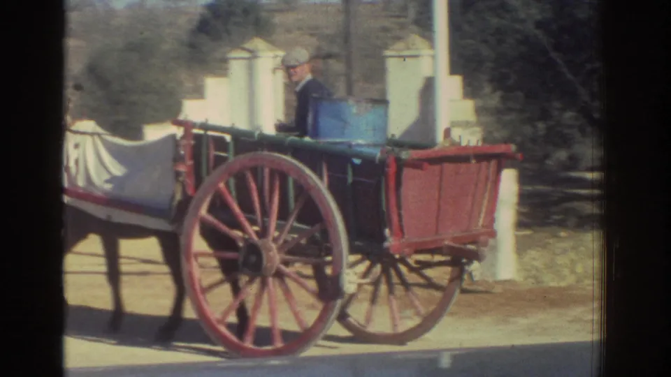 1976 Greece home-movie frame of a man in a light cap driving a horse-drawn wooden buggy with red wheels and a blue barrel.