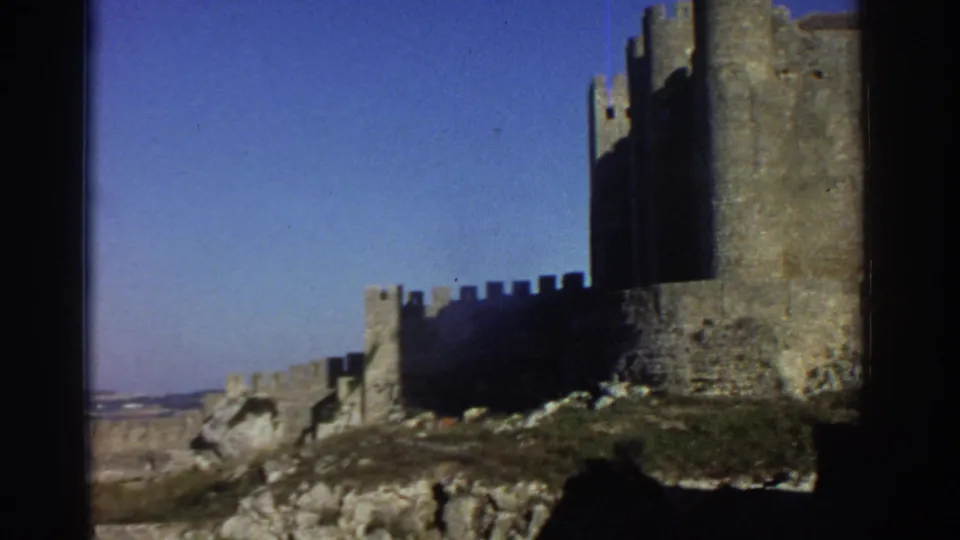 1976 Greece 8mm still of a castle wall and tower above rocky terrain, with distant countryside visible beyond.
