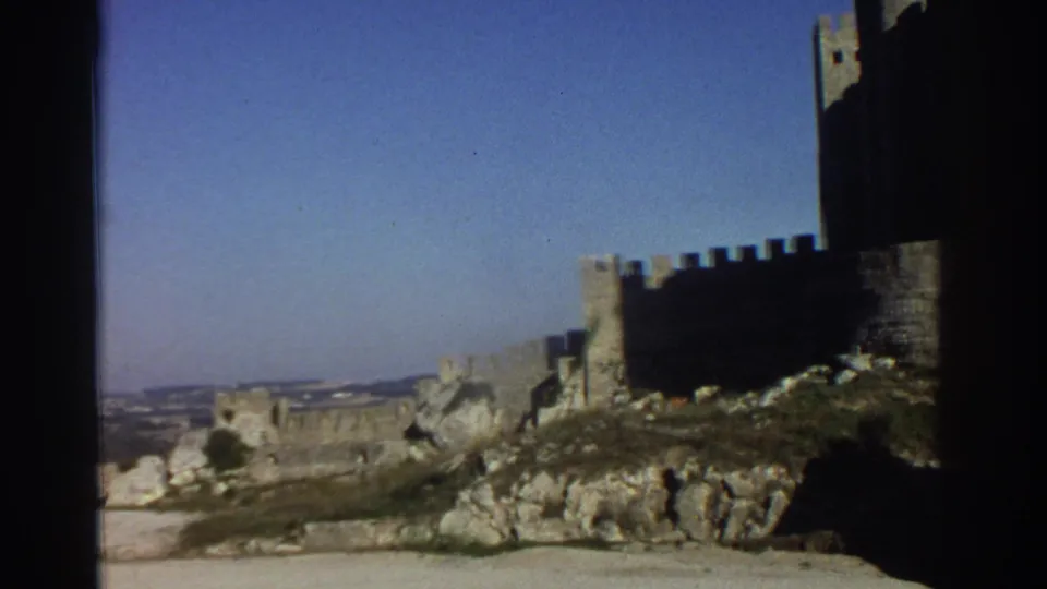 1976 Greece frame showing a crenellated castle wall on rocky terrain, with a distant landscape beyond the fortress.