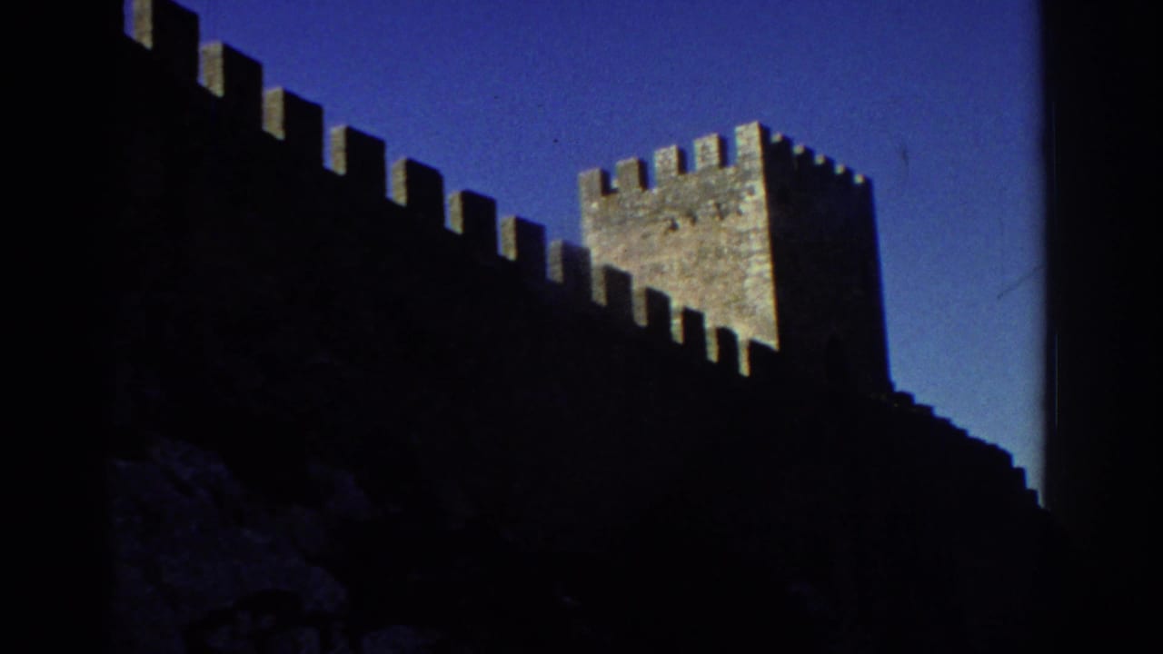 1976 Greece 8mm frame showing a dark stone fortress wall with battlements and a square tower under clear blue sky.