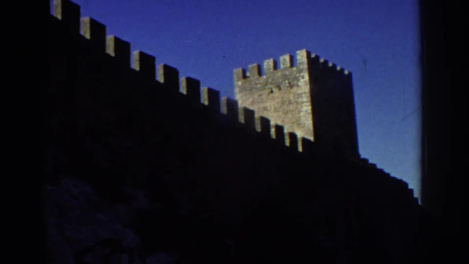 1976 Greece 8mm frame showing a dark stone fortress wall with battlements and a square tower under clear blue sky.