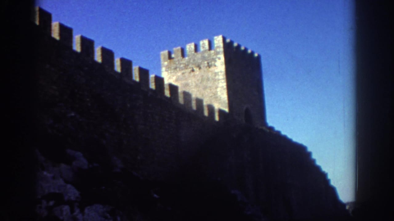 1976 Greece travel footage frame with crenellated fortress wall, square tower, and deep shadowed stonework beneath a blue sky.
