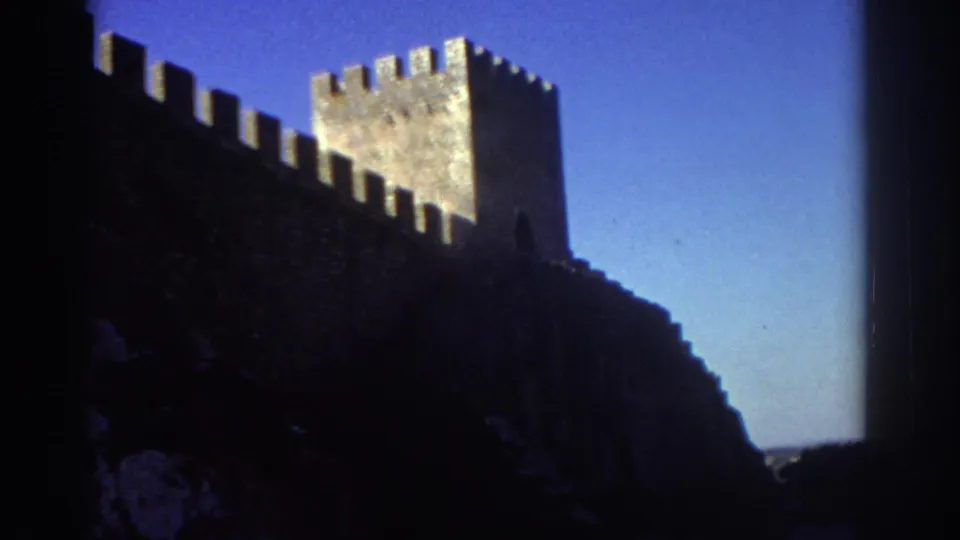 1976 Greece 8mm frame showing a crenellated stone wall and tower silhouetted above a rocky slope in bright daylight.