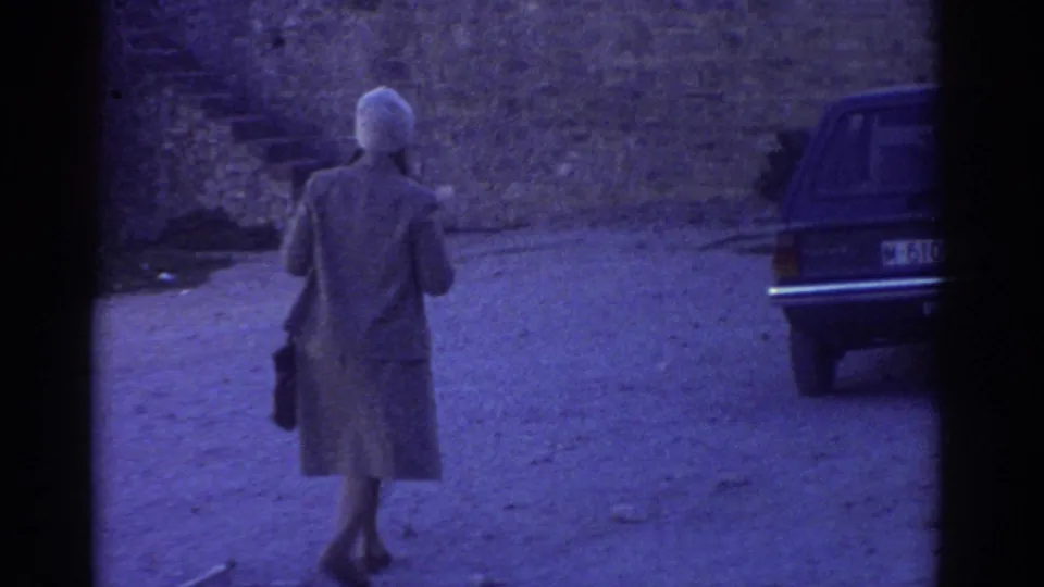 1976 Greece home-movie frame showing a woman in a light coat walking away near a parked car, with a stone fortress wall behind her.