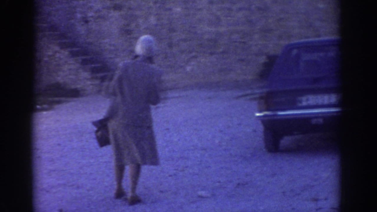 In 1976 Greece, a woman in a long coat and white head covering walks near a dark car beside a stone castle wall.