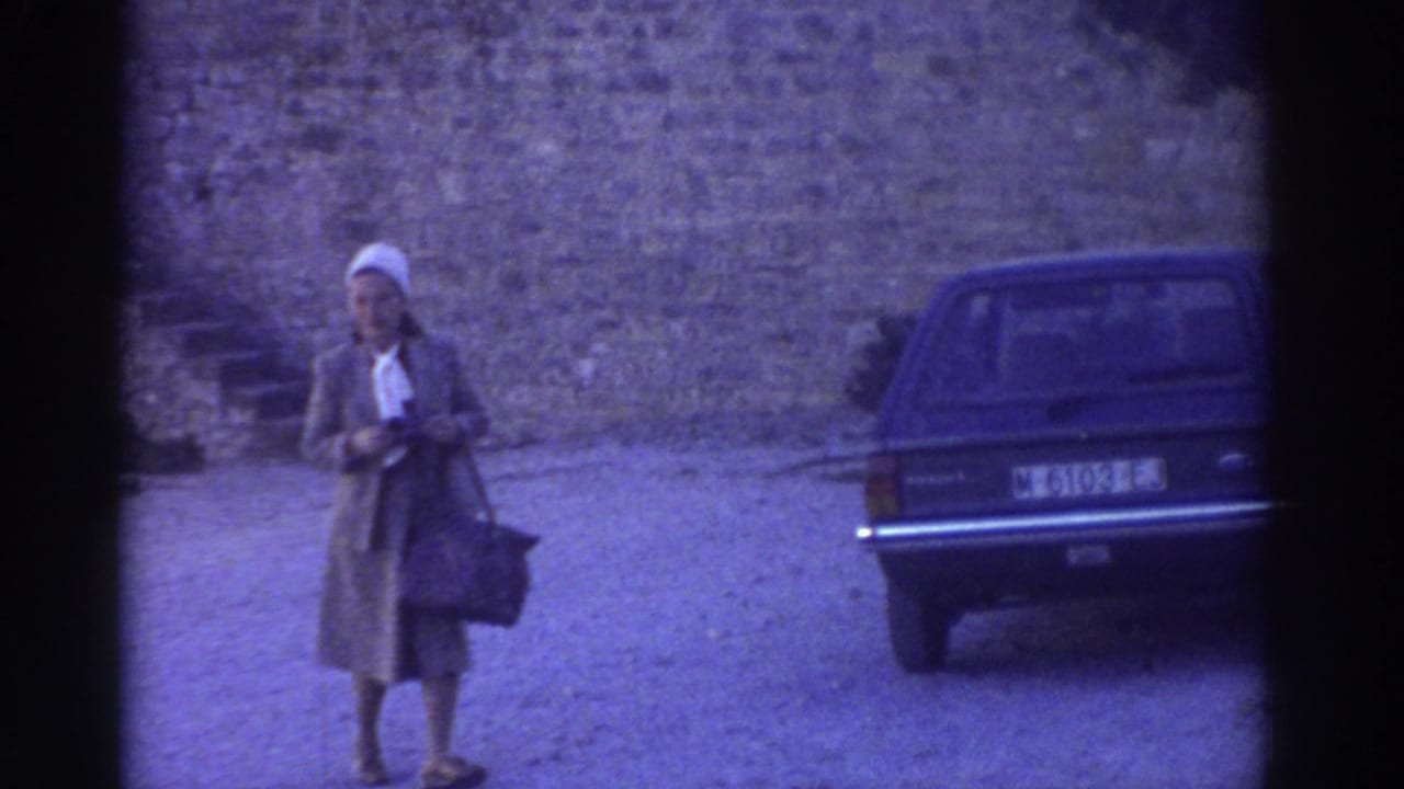 In 1976 Greece, a woman with a handbag stands or walks toward the camera near a dark car and castle wall.