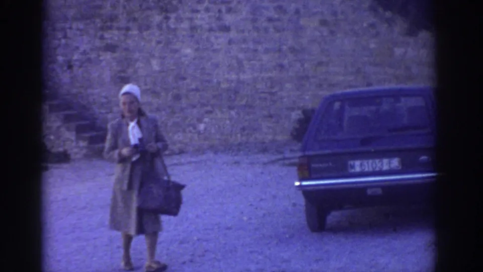 In 1976 Greece, a woman with a handbag stands or walks toward the camera near a dark car and castle wall.