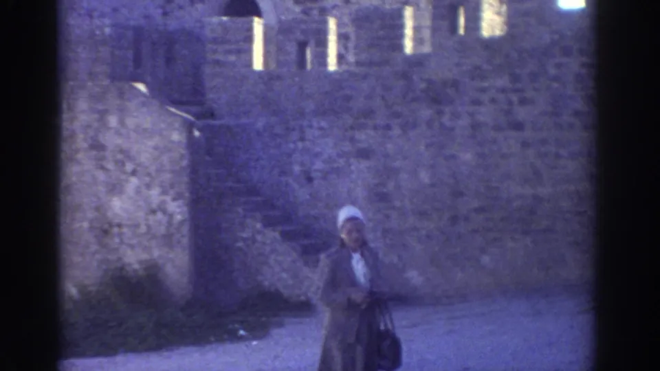 In 1976 Greece, the woman appears below stone castle walls, with steps and small wall openings visible behind her.