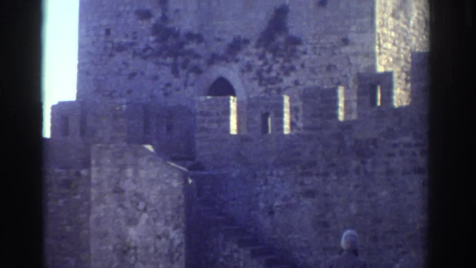 In 1976 Greece, the camera frames castle masonry, steps, narrow openings, and a partial view of the woman below.
