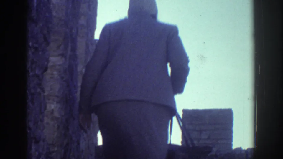 1976 Greece home-movie frame of a woman seen from behind climbing stone steps near an old masonry wall.