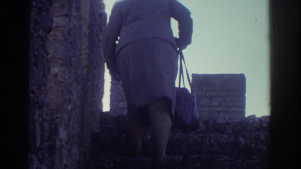 1976 Greece 8mm frame showing a woman in a coat and skirt carrying a handbag up stone steps by castle-like walls.